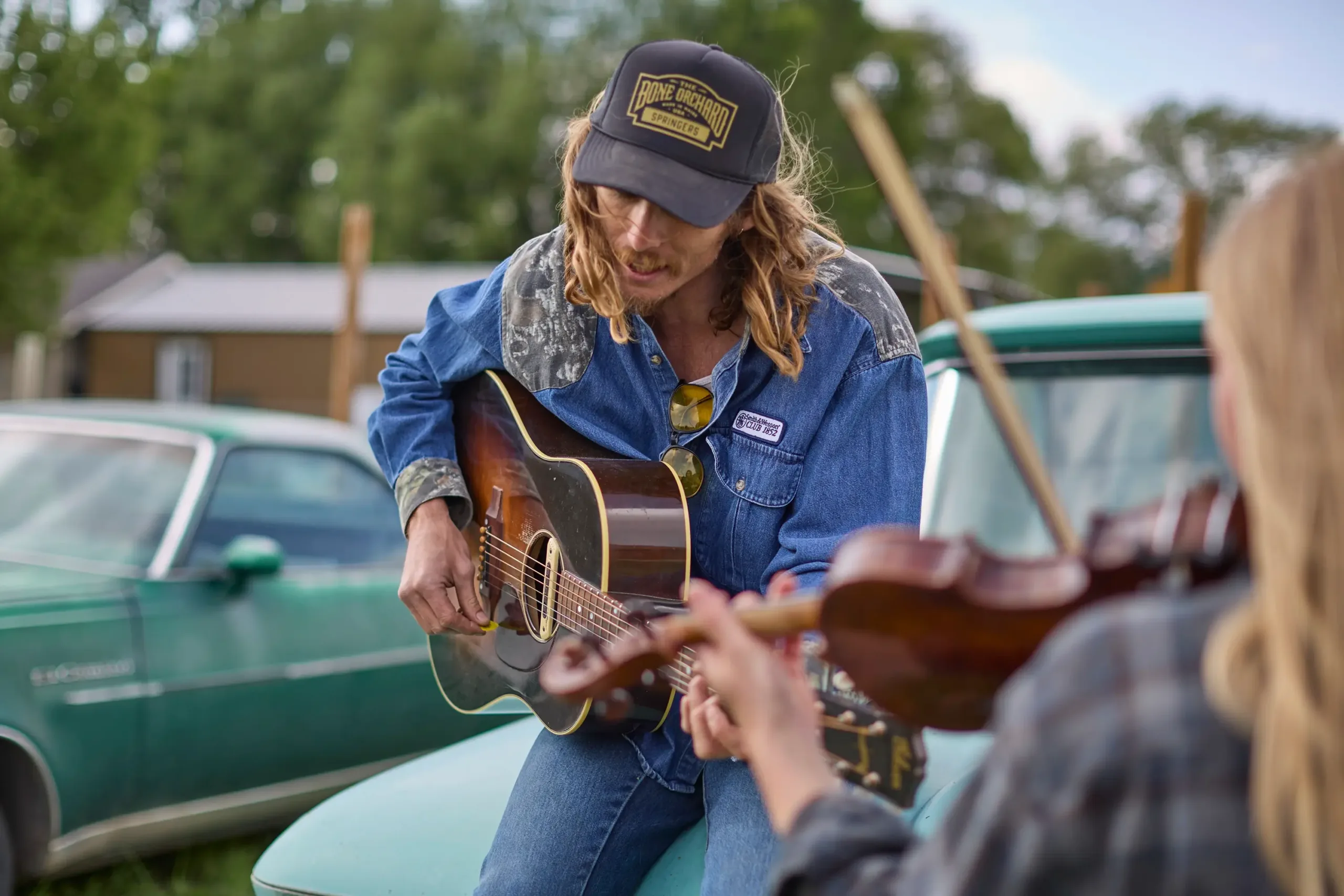 A man playing his guitar while sitting on a truck. A woman is in the foreground with her back to the camera